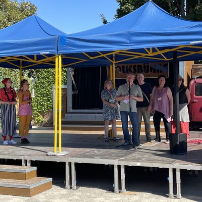 Bild vergr&ouml;&szlig;ern: Foto: Mehrere Menschen unter zwei blauen Pavillon-Zelten auf einer Bühne.