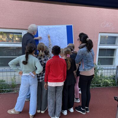 Bild vergr&ouml;&szlig;ern: Foto: Viele Kinder, ein Mann und eine Frau stehen um eine blaue Stellwand herum, an der ein Stadtplan hängt.