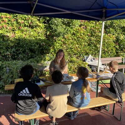 Bild vergr&ouml;&szlig;ern: Foto: Vier Kinder sitzen auf einer Bank. Gegenüber sitzt eine junge Frau.