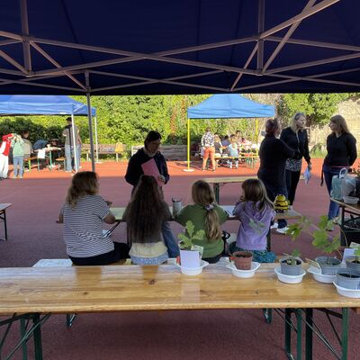 Bild vergr&ouml;&szlig;ern: Foto: Kinder sitzen auf einer Bank, Erwachsene stehen herum. Davor ein Tisch mit kleinen Setzlingen.