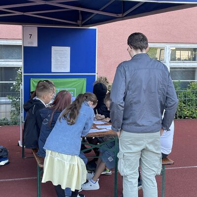 Bild vergr&ouml;&szlig;ern: Foto: Kinder sitzen an einem Tisch, ein Mann steht davor.