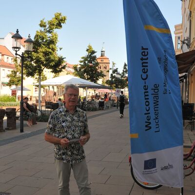 Foto: Ein Mann steht neben einer hellblauen Flagge in einer Fußgängerzone.