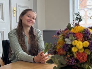Bild vergr&ouml;&szlig;ern: Eine junge Frau sitzt an einem Tisch und lächelt in die Kamera. Im Vordergrund ist ein Blumenstrauß zu sehen.