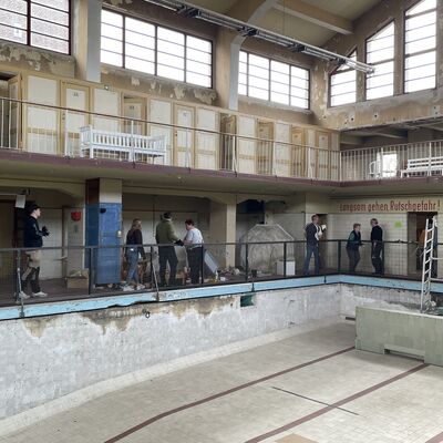 Bild vergr&ouml;&szlig;ern: Foto: Menschen laufen am Rand eines großen, leeren Schwimmbeckens herum.
