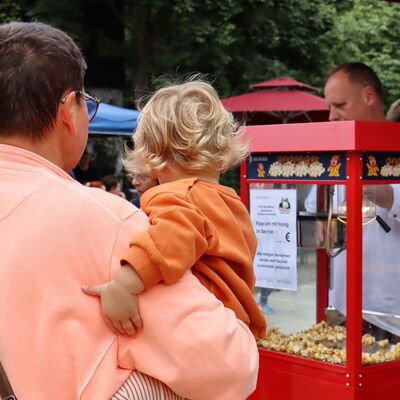 Bild vergr&ouml;&szlig;ern: Eine Frau hält ein kleines Kind auf dem Arm. Sie stehen mit dem Rücken zur Kamera an einem Popcornstand.