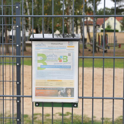 Foto: Ein Aushangkasten an einem Zaun. Im Hintergrund ein Spielplatz.