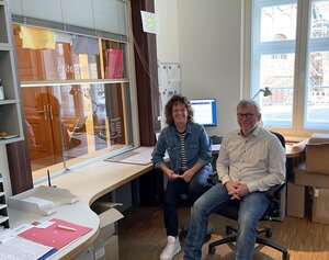 Bild vergr&ouml;&szlig;ern: Foto: Eine Frau und ein Mann sitzen in einem Büro mit einer großen Glasscheibe.