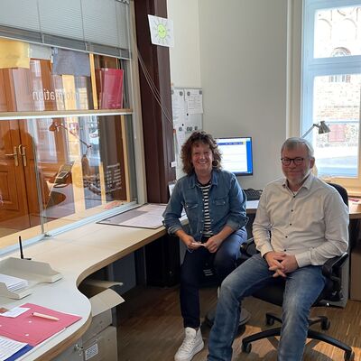 Foto: Eine Frau und ein Mann sitzen in einem Büro mit einer großen Glasscheibe.
