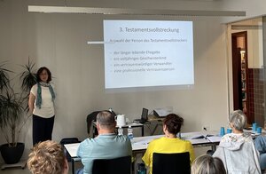 Bild vergr&ouml;&szlig;ern: Foto: Eine Frau steht vor einer Wand, auf die eine Präsentation projiziert wird. Vor ihr sitzen Menschen auf Stühlen.