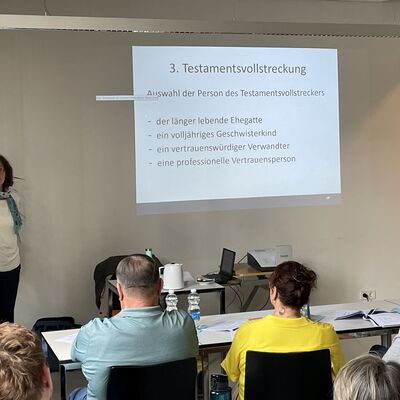 Foto: Eine Frau steht vor einer Wand, auf die eine Präsentation projiziert wird. Vor ihr sitzen Menschen auf Stühlen. 