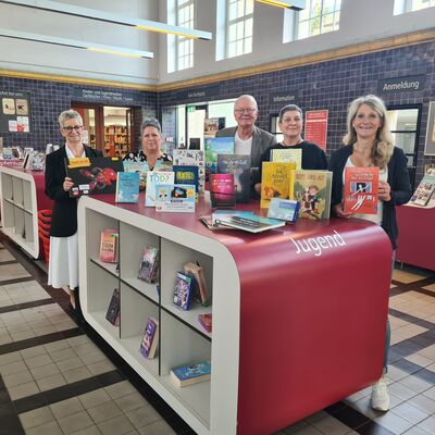 Foto: Vier Frauen und ein Mann stehen in einer großen Halle hinter einem weiß-roten Regal mit Büchern.