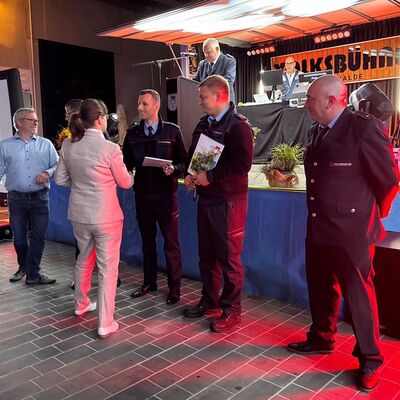 Vor einer Bühne stehen Feuerwehrleute und bekommen Urkunden überreicht.
