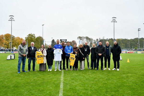 Bild vergr&ouml;&szlig;ern: Mehrere Menschen stehen auf einem Fußballplatz und lächeln in die Kamera.