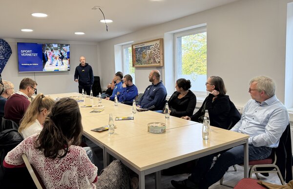 Bild vergr&ouml;&szlig;ern: Menschen sitzen um einen Tisch und schauen zu einem Bildschirm.