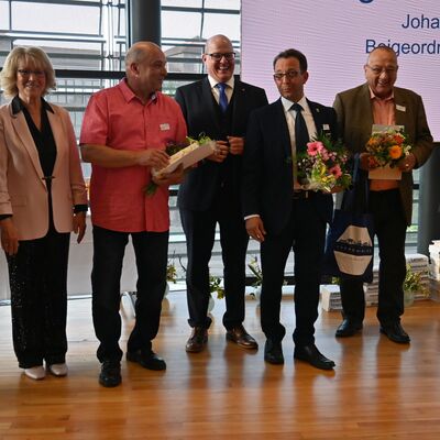 Mehrere Personen stehen auf einer Bühne, einige halten Blumen und Präsente in den Händen.