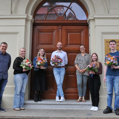 Foto: Drei Männer und vier Frauen stehen auf einer Treppe vor einer großen Tür.